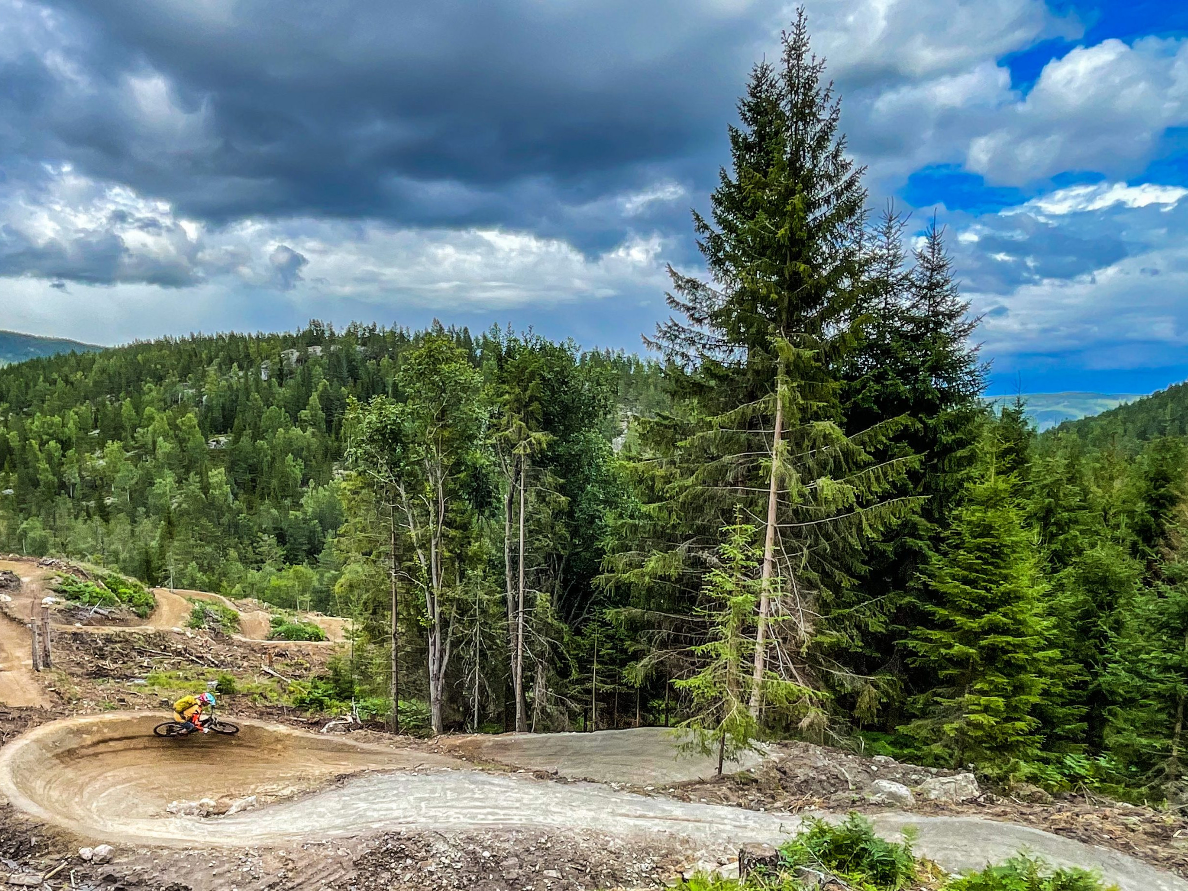 Cyclist rides through forested mountain trail with dramatic clouds overhead. Explore 
