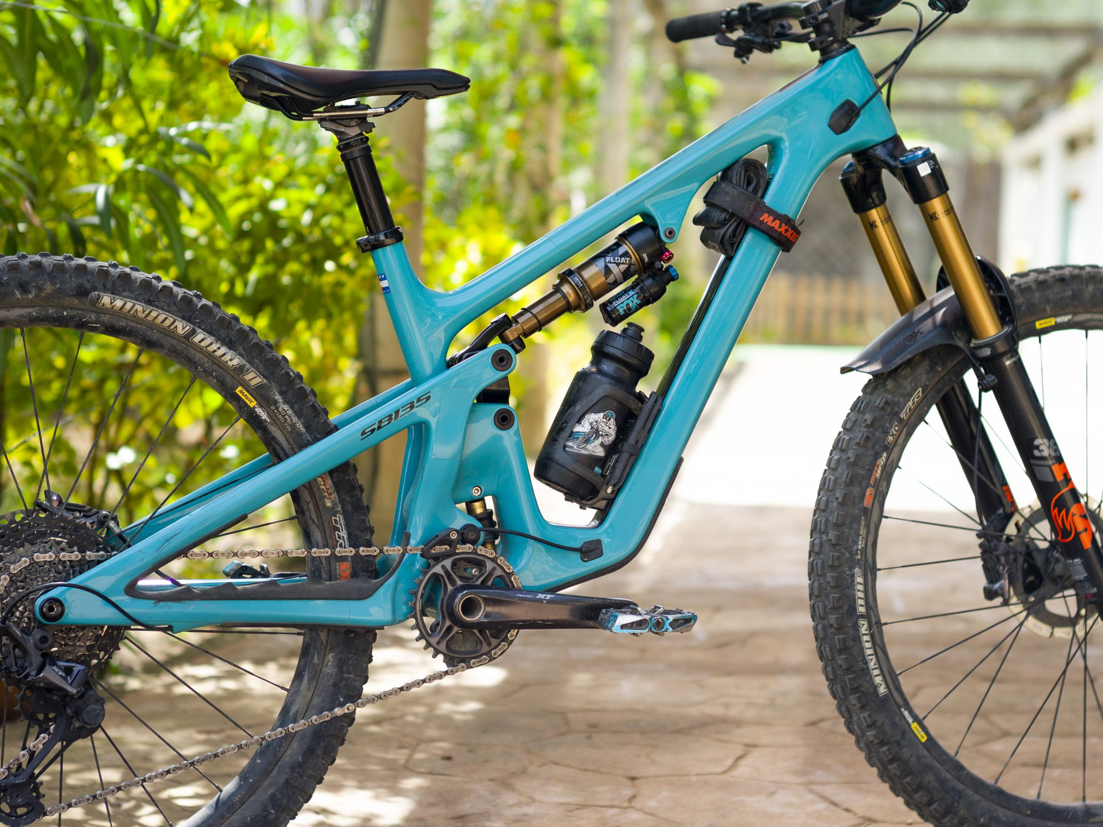 Blue high-performance mountain bike, full-suspension design, equipped with Maxxis tires and water bottle, parked outdoors in a garden setting. Mountain Bike United.