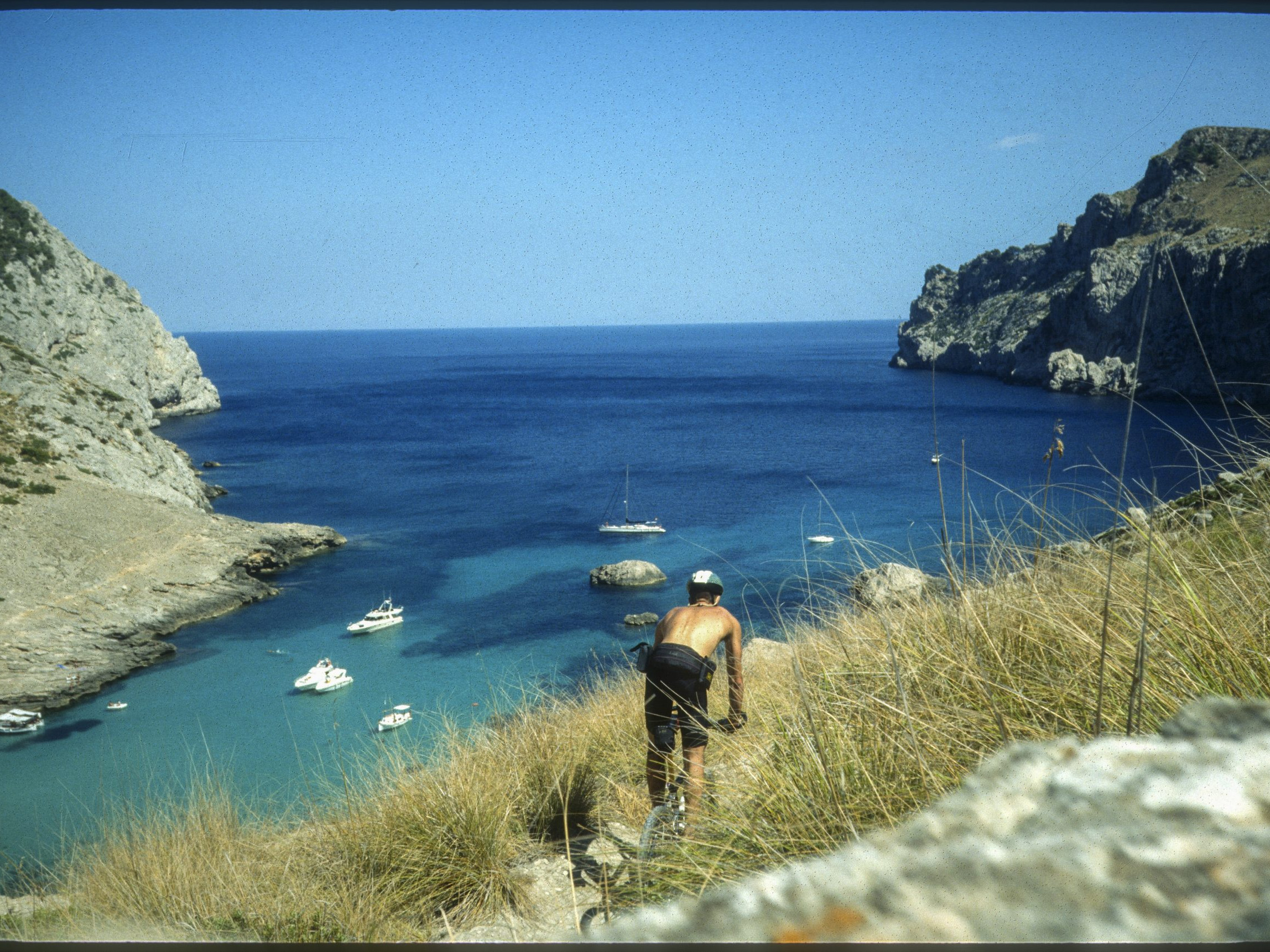 Cyclist descends a rocky trail towards a scenic coastal bay with boats in clear blue water, surrounded by cliffs in a sunny Mediterranean landscape. Mountain Bike United.