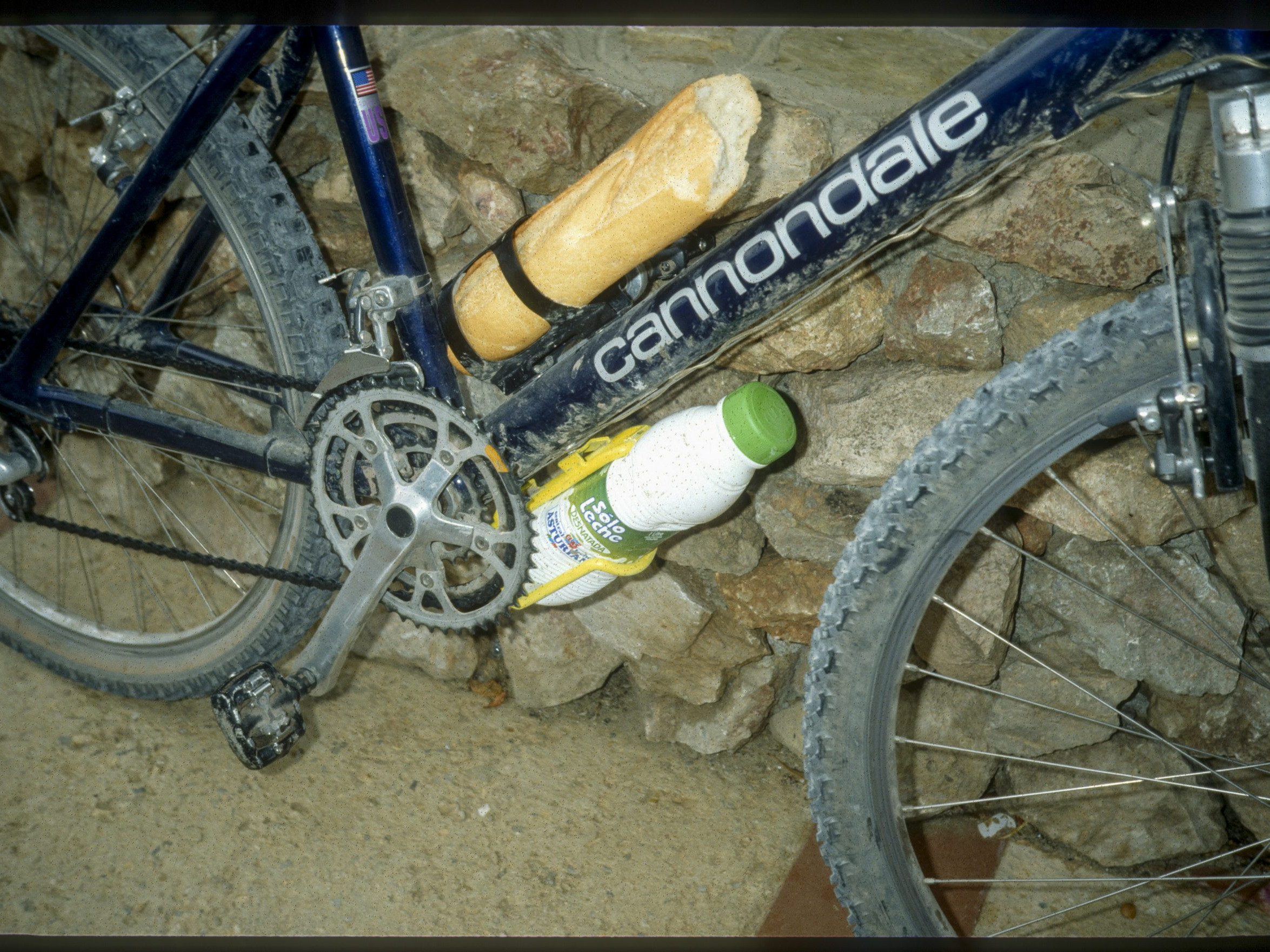 Cannondale bicycle with baguette and bottle of lemon soda on frame, parked against stone wall. Mountain bike, outdoor adventure, cycling gear. Mountain Bike United.