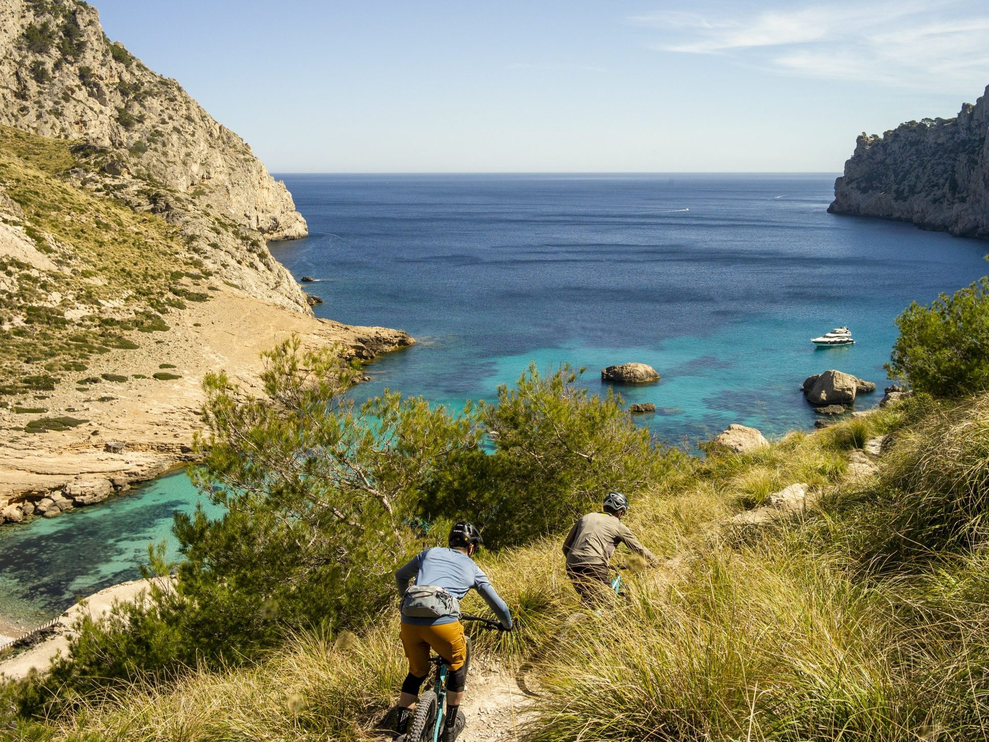 Cyclists biking down a coastal trail with lush cliffs and turquoise waters, and a yacht floating in the Mediterranean Sea in the background. Mountain Bike United.