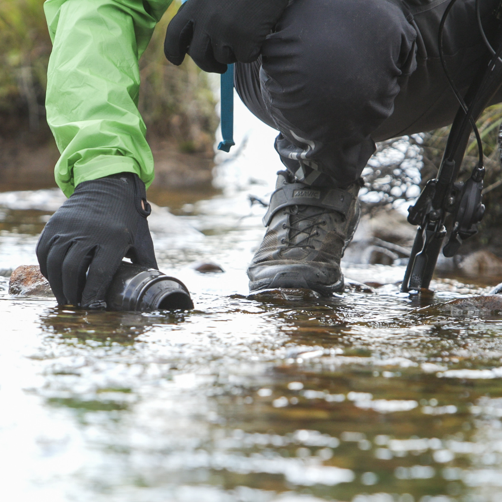 Cyclist in green jacket kneeling by stream, filling cup with water. Dan Milner, A Single Drop concept in outdoor adventure. Mountain Bike United.