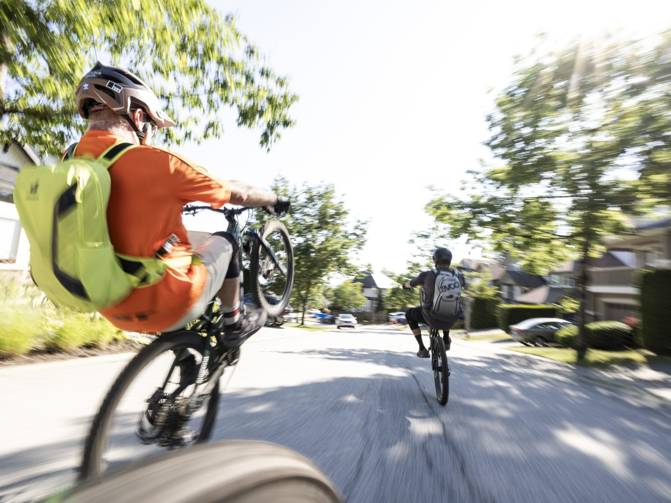 Two cyclists perform wheelies on a sunny street, showcasing bike skills. Hans Rey inspired action, capturing urban cycling energy and excitement. Mountain Bike United.