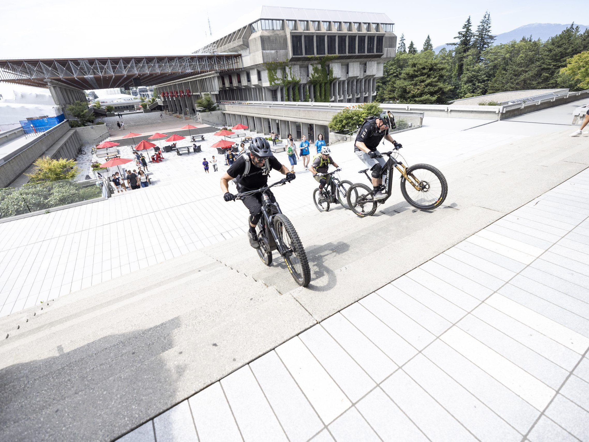 Cyclists ride up steps at a modern campus, showcasing mountain biking skills. Possible inclusion of Hans Rey for biking techniques. Mountain Bike United.