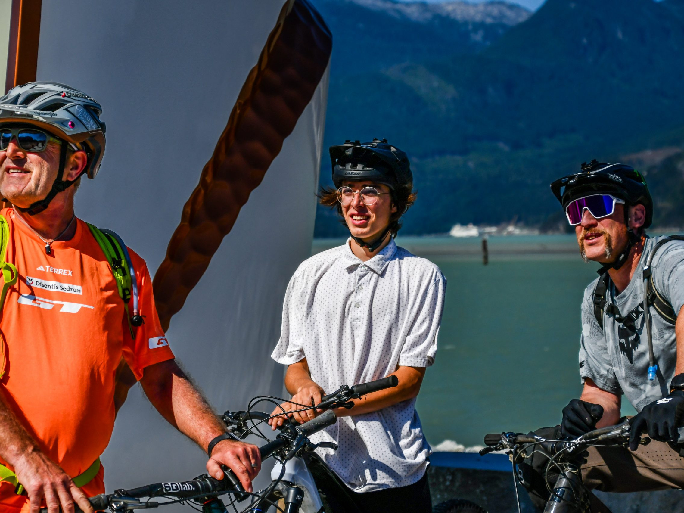Three cyclists with helmets enjoying a scenic mountain view near water. Ideal for terms: mountain biking, cyclists, outdoor adventure, Hans Rey. Mountain Bike United.