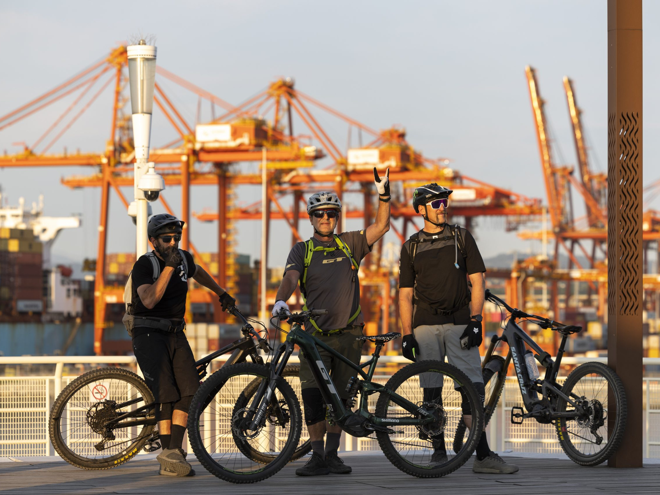 Three cyclists with mountain bikes at a port with cranes in the background, possibly featuring Hans Rey, emphasizing adventure and cycling. Mountain Bike United.