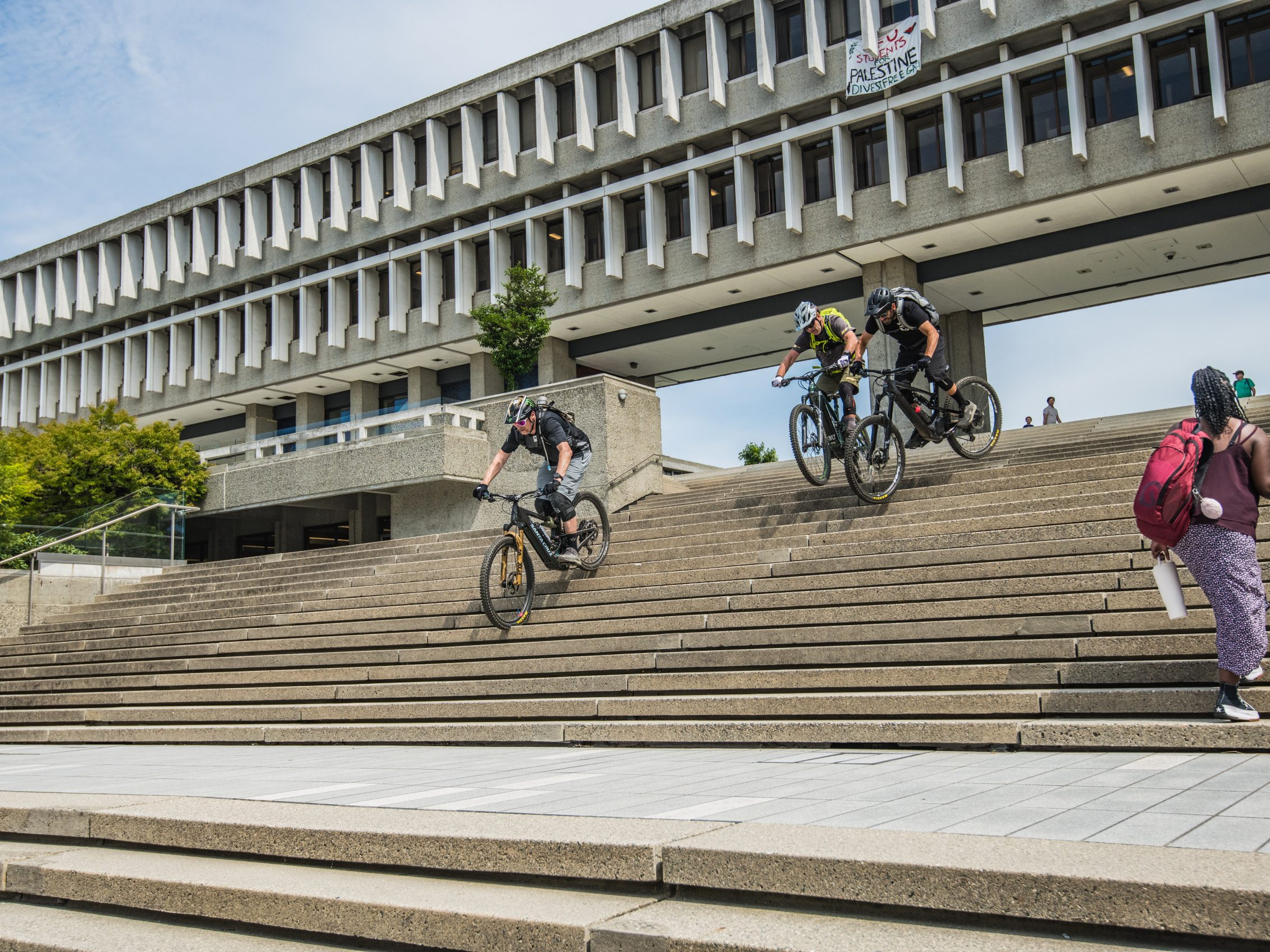 Mountain bikers ride down concrete steps in front of a modern building. Action, cycling, urban biking. Hans Rey inspiration. Mountain Bike United.