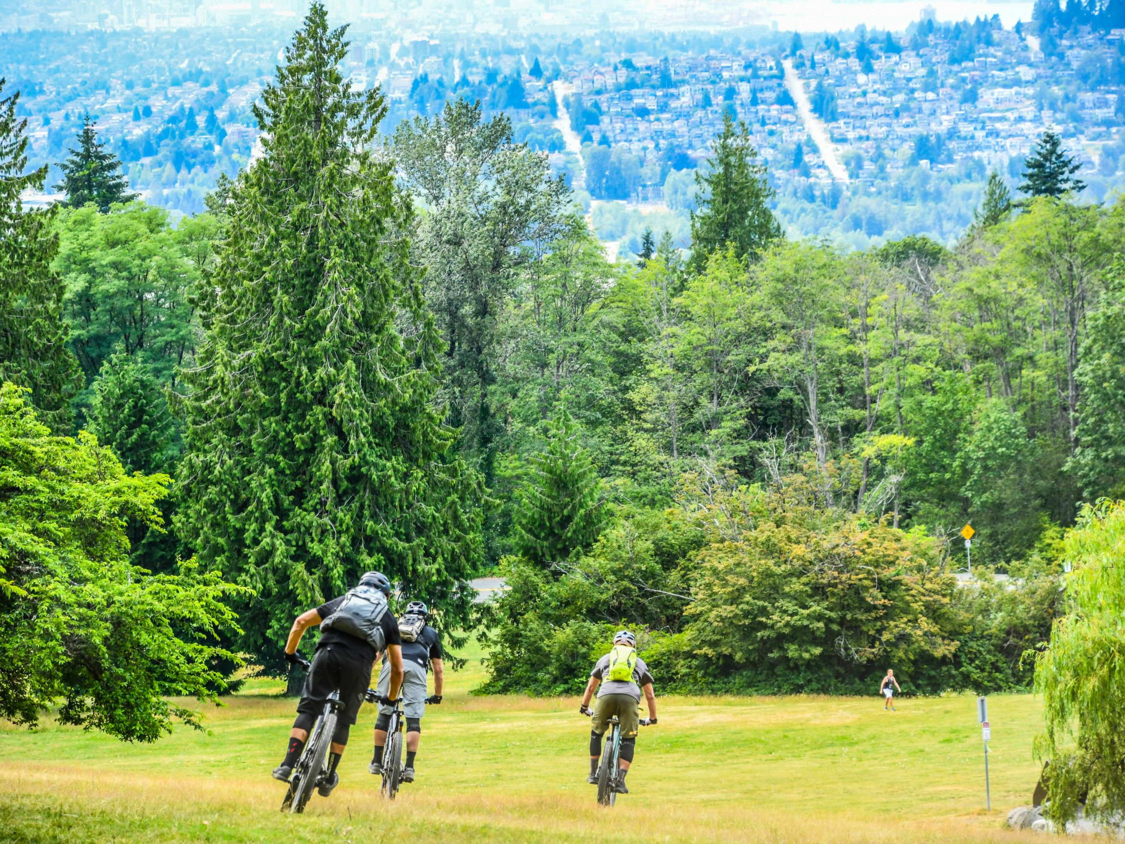 Group of mountain bikers riding downhill through a forest, exploring trails. Greenery surrounds them with a city view in the background. Mountain Bike United.