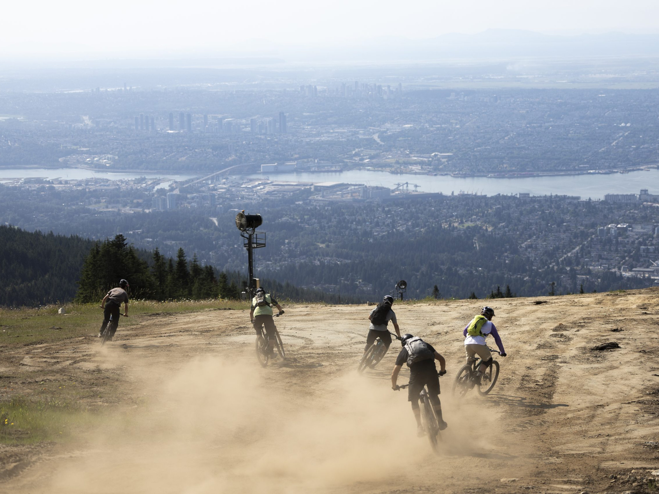 Mountain bikers ride downhill on a dusty trail with cityscape views, related to Hans Rey adventure cycling and scenic biking routes. Mountain Bike United.