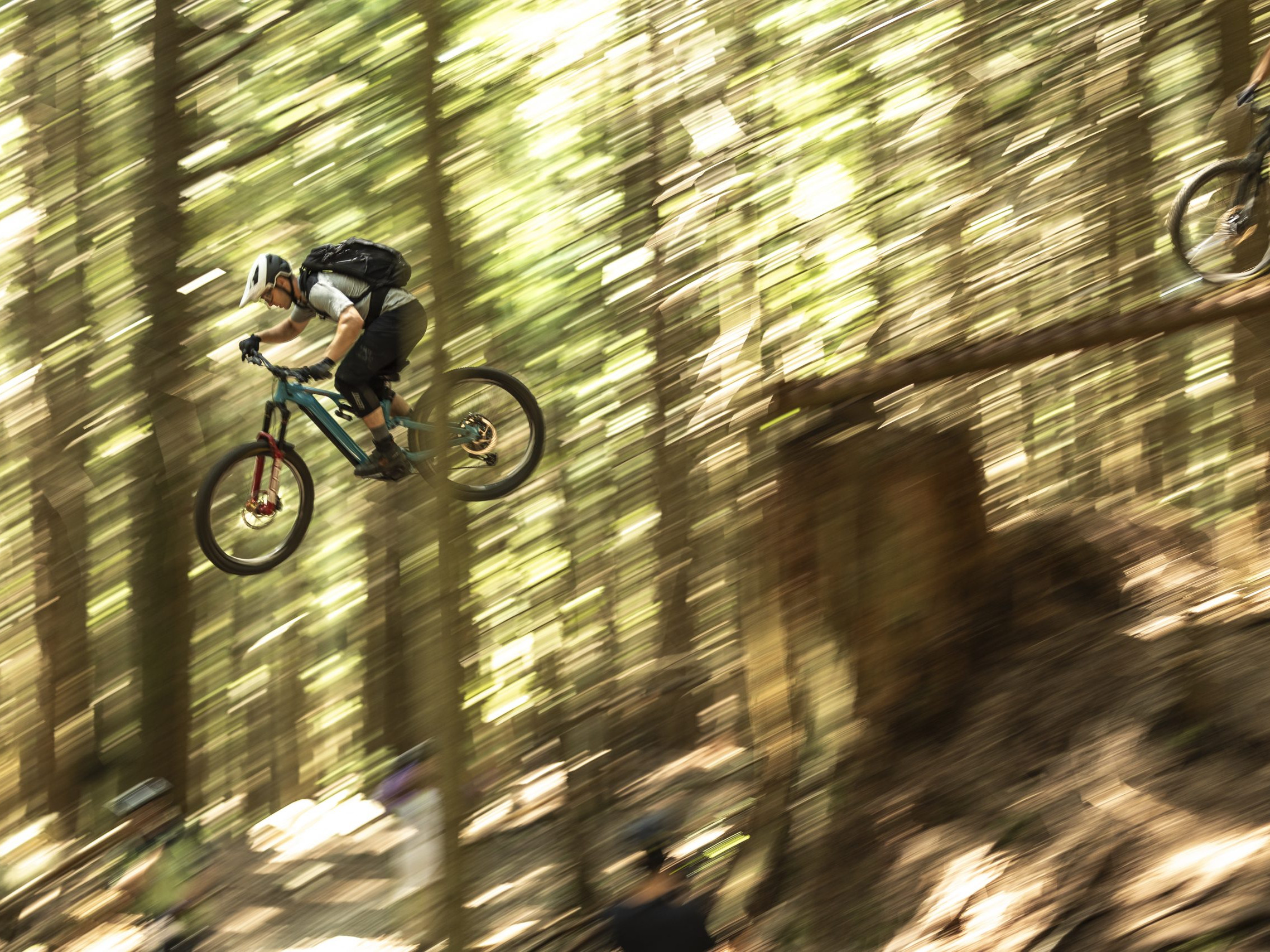 Mountain biker performing a high jump in a dense forest, showcasing thrilling outdoor adventure sports similar to Hans Rey's biking stunts. Mountain Bike United.