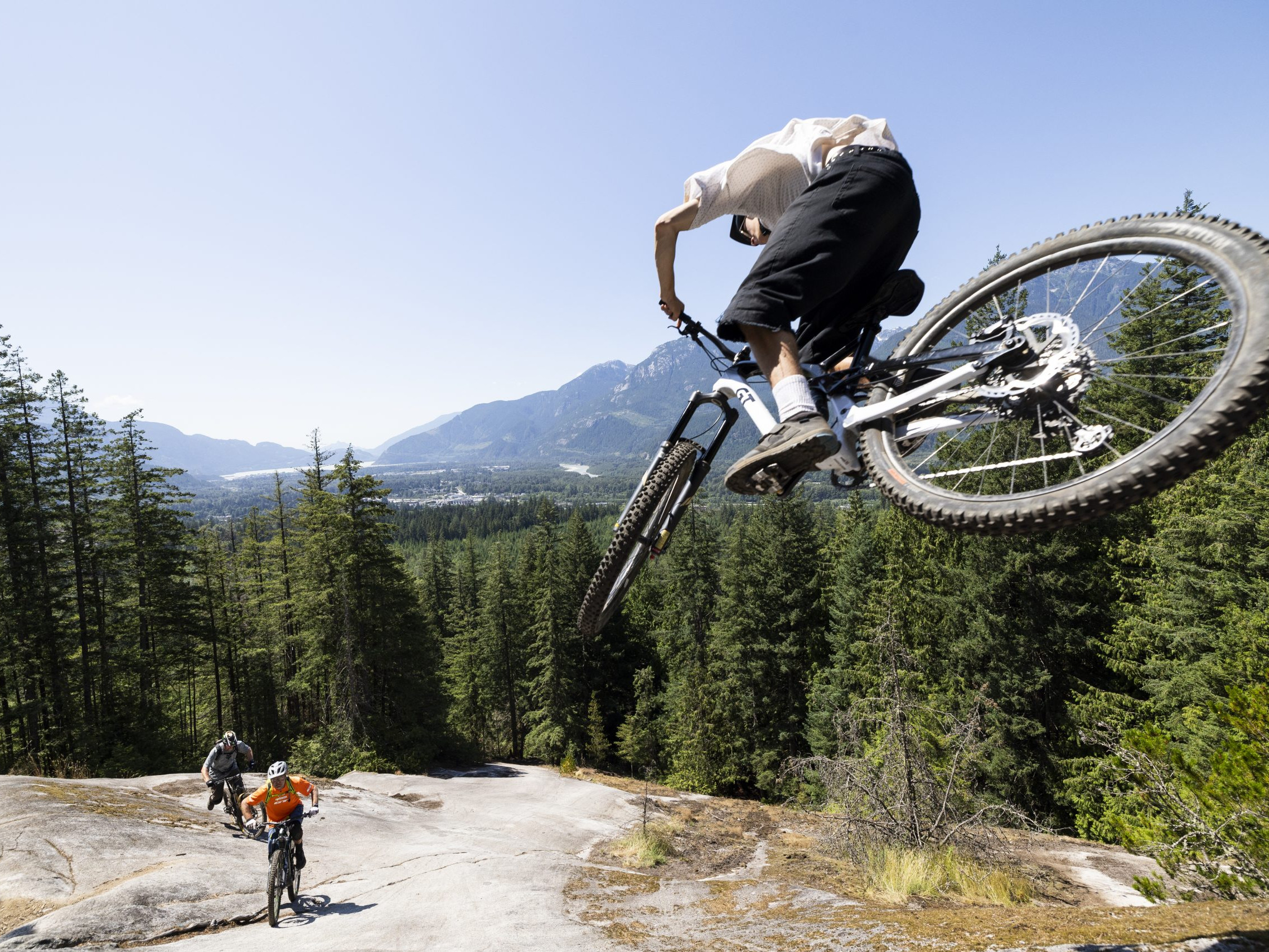 Mountain biker performing jump on trail with scenic forest and mountains, possibly Hans Rey. Action sports, adventure biking, outdoor exploration. Mountain Bike United.