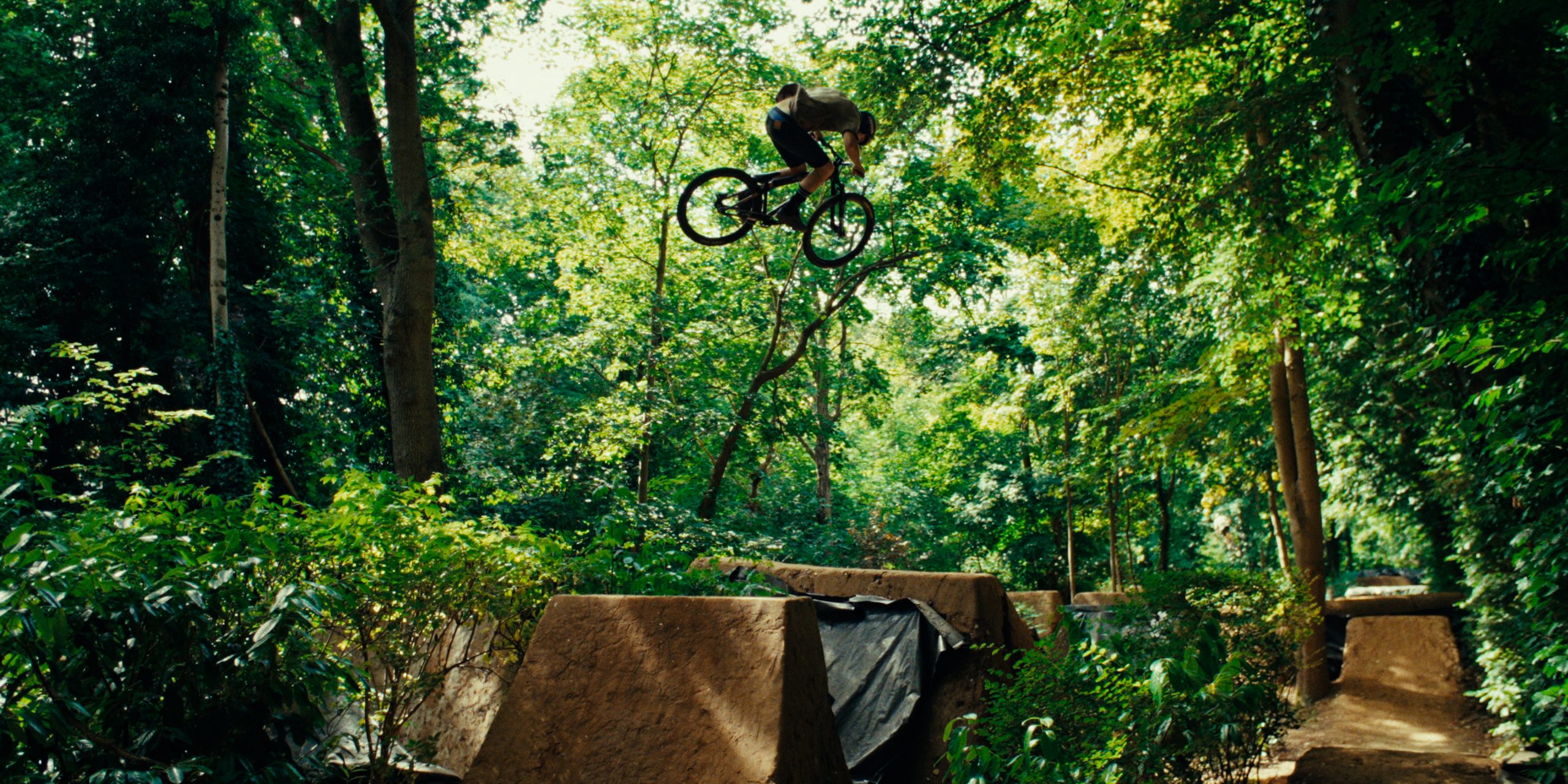 Mountain biker performing aerial stunt over dirt jump in lush forest setting, showcasing extreme sports and outdoor adventure. Mountain Bike United.