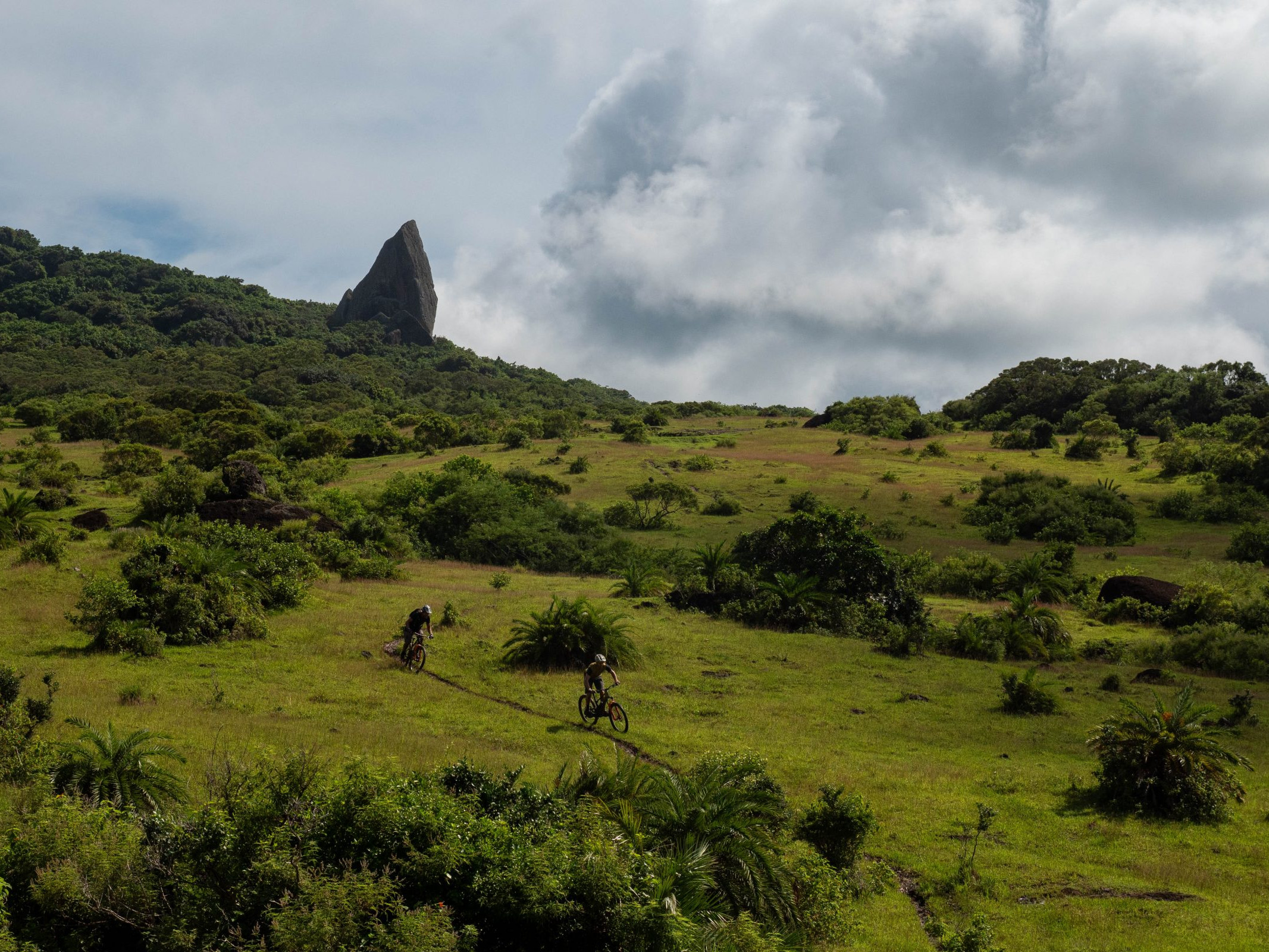 Cyclists on a scenic trail with lush greenery and a rocky peak in the backdrop; related to Andrew Neethling Lifting The Lid. Mountain Bike United.